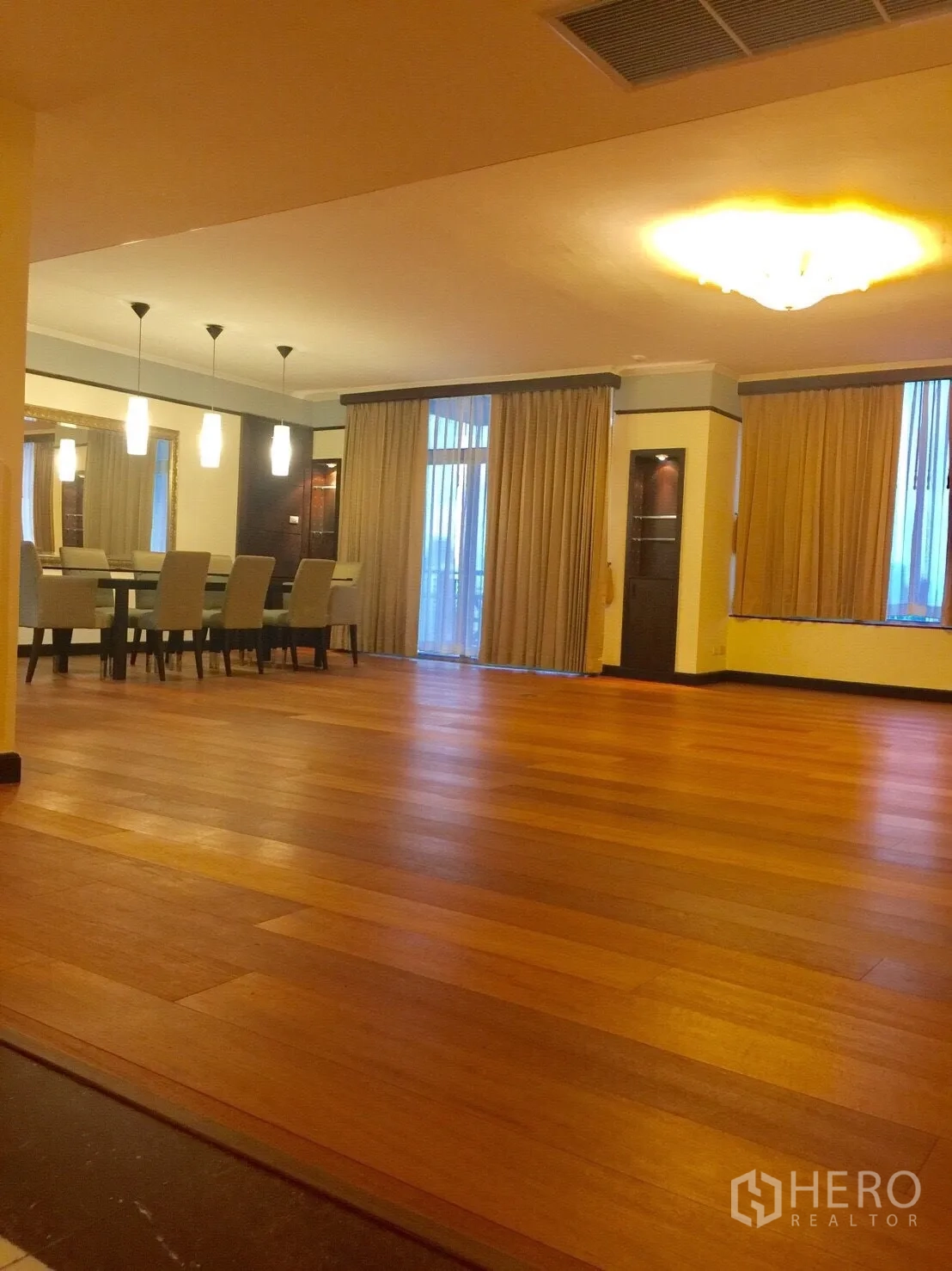 Condominium for rent in Pathum Wan, Bangkok - Spacious open-plan living and dining area with warm wood floors, pendant lights and floor-to-ceiling curtains opening to a balcony.