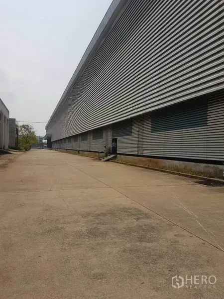 工厂 出租在Mueang Samut Sakhon, Samut Sakhon - 宽阔混凝土车道与长条形金属外墙的工厂外观，设多处装卸区，位于Samut Sakhon。