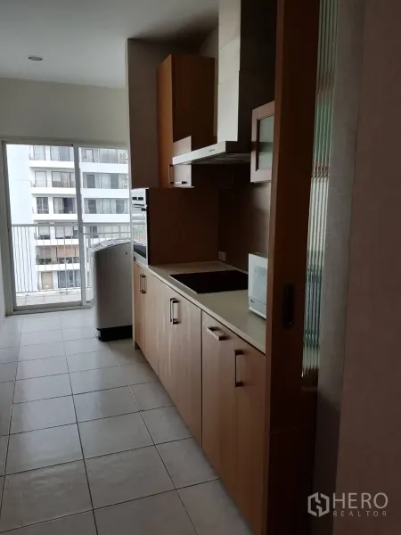 Condominium for rent or sale in Watthana, Bangkok - Kitchen view toward the balcony with cooktop, wood cabinets, and city outlook through sliding doors.