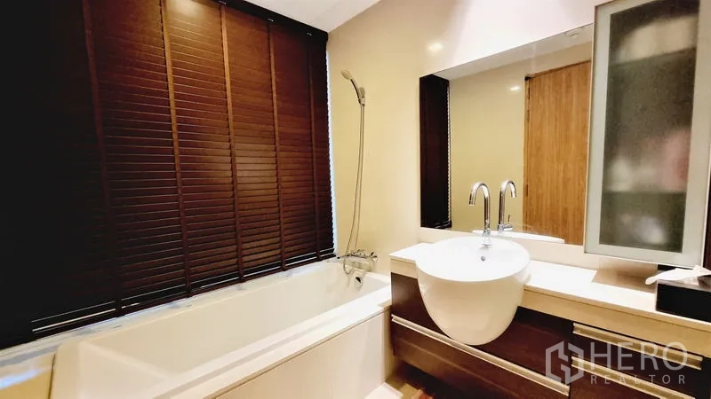 Condominium for sale in Watthana, Bangkok - Modern bathroom with bathtub, handheld shower, round basin and large mirror with dark blinds.