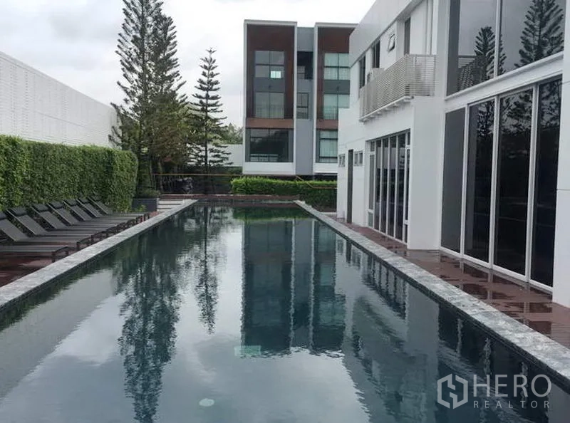 Townhouse for rent in Suan Luang, Bangkok - Long outdoor swimming pool with sun loungers next to a glass-fronted clubhouse.