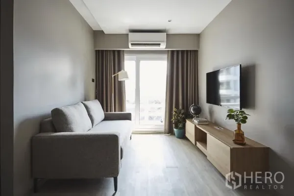 Apartment for rent in Khlong Toei, Bangkok - Bright living room with grey sofa, wall-mounted TV, balcony doors and air conditioner.