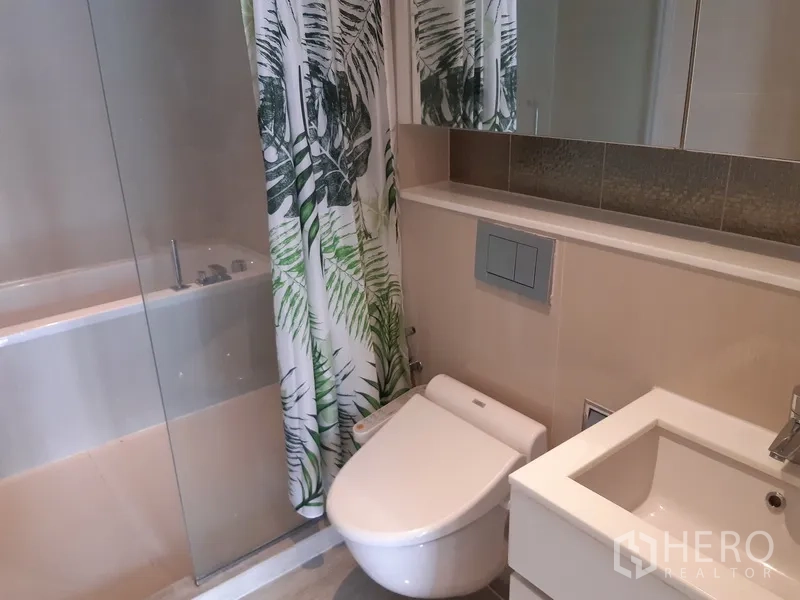 Condominium for rent in Watthana, Bangkok - Modern bathroom with bidet toilet, vanity sink, and leafy shower curtain beside a bathtub.