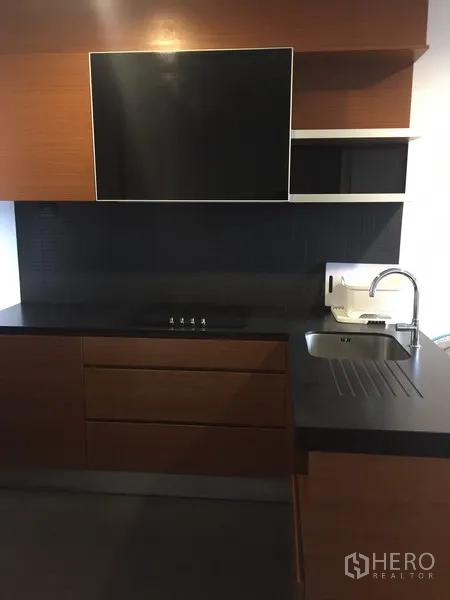 Condominium for rent in Khlong Toei, Bangkok - Modern kitchenette with dark countertop, sink, induction hob and wood cabinetry.