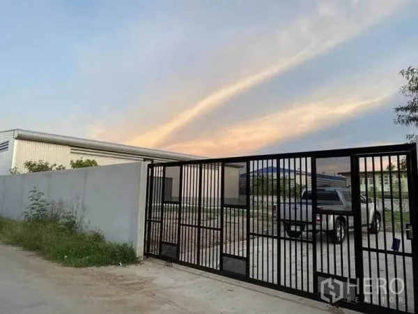 Warehouse for rent in Sam Phran, Nakhon Pathom - Secure black sliding gate and concrete wall leading to a modern steel warehouse yard at sunset in Sam Phran.