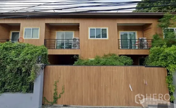 Townhouse for rent or sale in Watthana, Bangkok - Two-storey modern townhouse exterior with warm wood cladding, twin balconies, sliding glass doors, black metal railings, gated wall and lush greenery in the Sukhumvit–Ekkamai area.