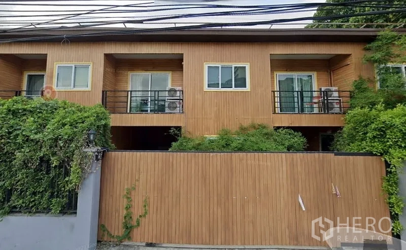 Townhouse for rent or sale in Watthana, Bangkok - Two-storey modern townhouse exterior with warm wood cladding, twin balconies, sliding glass doors, black metal railings, gated wall and lush greenery in the Sukhumvit–Ekkamai area.