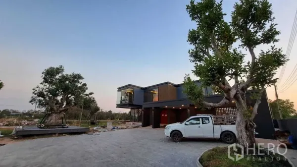 บ้าน ขายในChachoengsao - บ้านเดี่ยวสองชั้นสไตล์โมเดิร์นยามเย็น มีกระจกมุมแคนทิลีเวอร์ ลานจอดรถกว้าง ต้นไม้ใหญ่ และรถกระบะจอดหน้าบ้าน