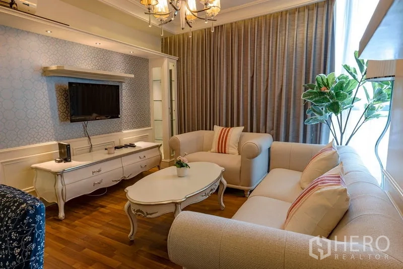 Condominium for rent in Watthana, Bangkok - Living room with cream sofas, coffee table, TV console and chandelier by a tall window and indoor plant.