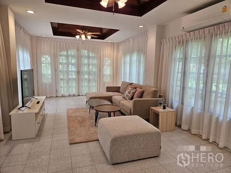 Detached House for rent - Sunlit lounge with L-shaped beige sofa, tiled floor, and arched window wall.