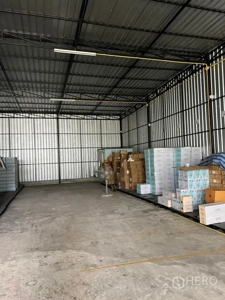 货栈 出租在Lat Phrao, Bangkok - 宽阔通道的金属屋顶仓库，混凝土地面、日光灯照明，托盘纸箱与立式风扇可见。