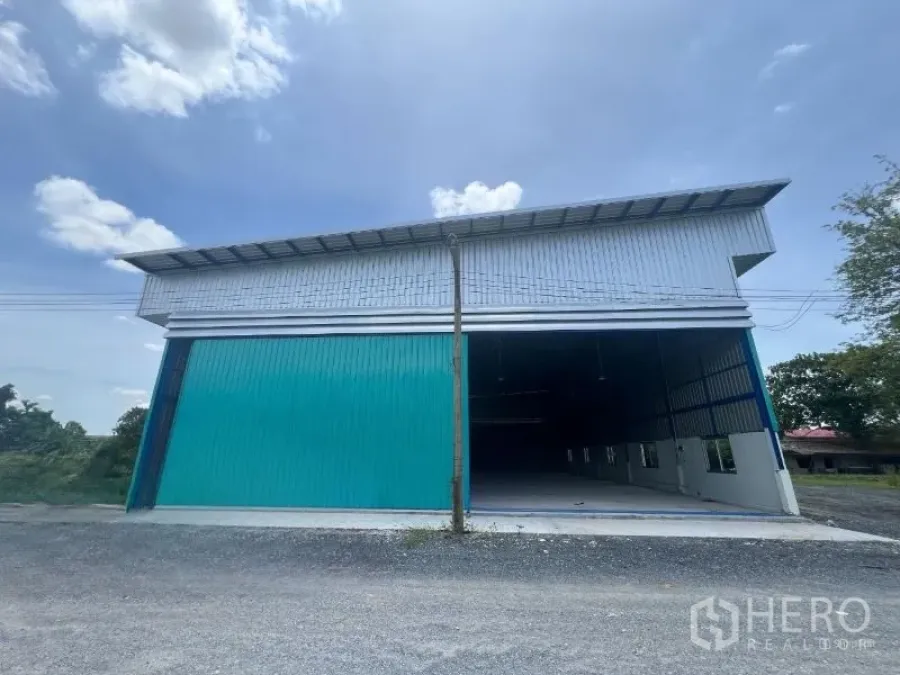 Warehouse for rent in Min Buri, Bangkok - Front view of a modern steel warehouse with teal sliding doors and a high roof along a gravel access road in Min Buri, Bangkok.