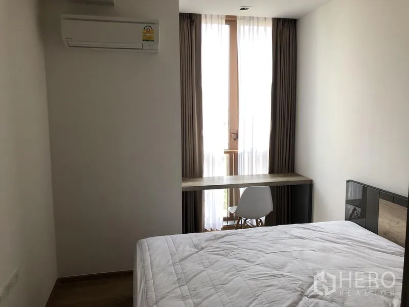 Condominium for rent in Watthana, Bangkok - Compact bedroom with air conditioning, bed, and a work desk by a tall curtained window.