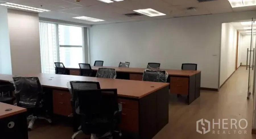 Office Space for rent in Bang Rak, Bangkok - Open-plan office with long workstations, swivel chairs, wood flooring, large windows with blinds, and recessed lighting near a glass entry.