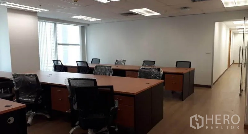 Office Space for rent in Bang Rak, Bangkok - Open-plan office with long workstations, swivel chairs, wood flooring, large windows with blinds, and recessed lighting near a glass entry.