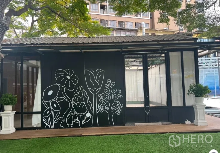 零售空间 出租在Watthana, Bangkok - 临街玻璃店面，黑色花卉墙、绿植花盆与草坪，适合咖啡店或精品店。