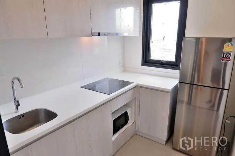 Condominium for rent in Huai Khwang, Bangkok - Compact white kitchen with electric cooktop, microwave, and stainless refrigerator.
