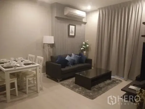 Condominium for rent in Huai Khwang, Bangkok - Cozy living room with gray sofa, coffee table, rug, and a compact dining nook under soft lighting.