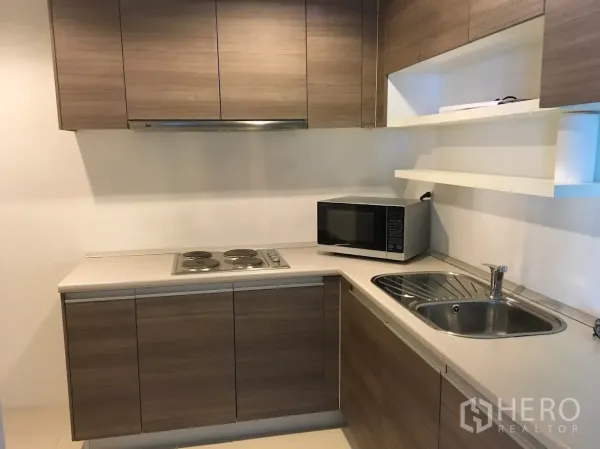 Condominium for rent in Huai Khwang, Bangkok - Modern L-shaped kitchen with wood cabinets, electric cooktop, microwave and stainless sink.