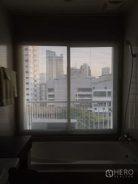 Condominium for rent in Watthana, Bangkok - Bathroom window with blinds overlooking the city skyline above a bathtub.