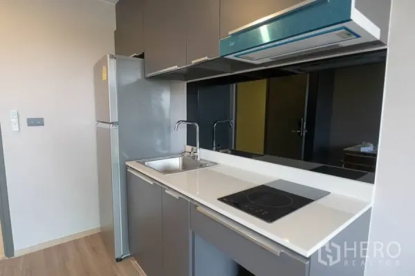 Condominium for rent or sale in Watthana, Bangkok - Compact kitchenette with grey cabinets, black glass backsplash, electric cooktop, hood, sink, and full-size fridge.