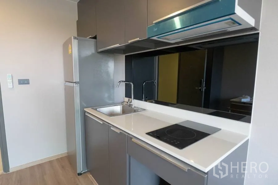 Condominium for rent or sale in Watthana, Bangkok - Compact kitchenette with grey cabinets, black glass backsplash, electric cooktop, hood, sink, and full-size fridge.