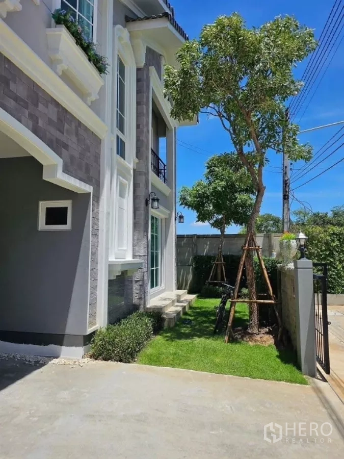 Detached House for rent in Thanyaburi, Pathum Thani - Side yard with grass lawn, steps to a sliding door and parking area beside the boundary wall.
