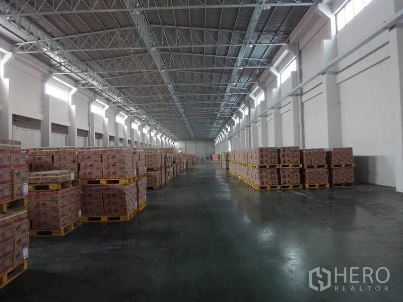 货栈 出租在Bang Sao Thong, Samut Prakan - 高跨仓库内部，两侧堆放托盘纸箱，钢桁架屋顶与天窗照明。