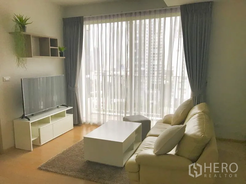 Condominium for rent in Watthana, Bangkok - Cozy living room with sofa, coffee table, and floor‑to‑ceiling window dressed with sheer curtains.