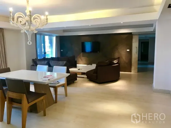 Condominium for rent or sale in Watthana, Bangkok - Spacious lounge with wall-mounted TV, sectional sofa, and dining table near the balcony doors.