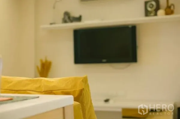 คอนโด ขายในWatthana, Bangkok - มุมห้องนั่งเล่นทีวีติดผนังและชั้นวางของ มีผ้าคลุมโซฟาสีเหลืองด้านหน้า.