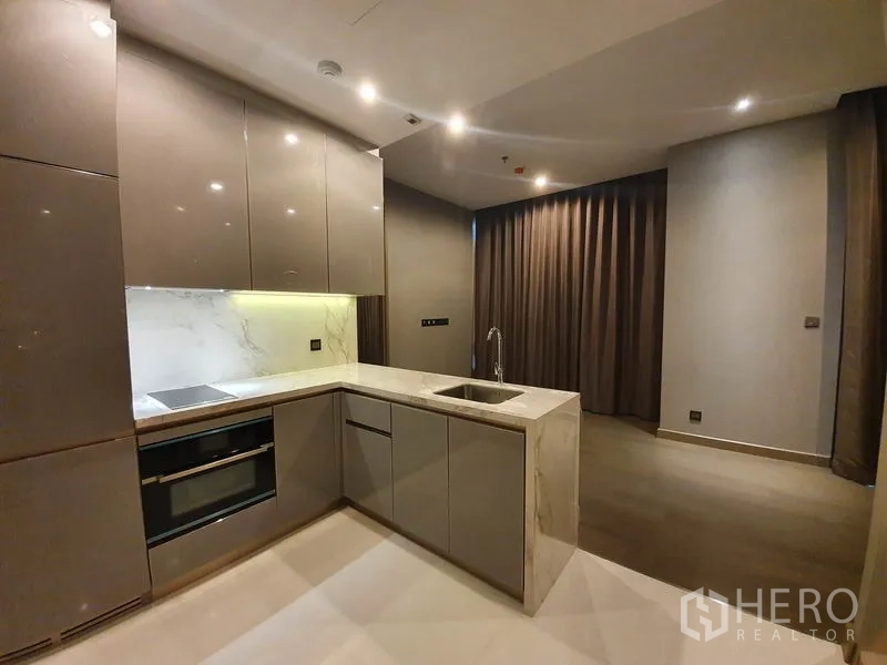 Condominium for rent in Huai Khwang, Bangkok - Modern L-shaped kitchen with marble counters, glossy cabinetry, built-in hob and oven, and sink beside a curtained living area.