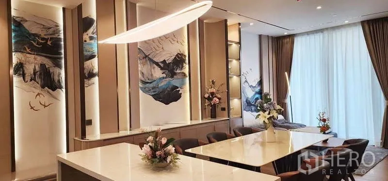 คอนโด ให้เช่า - โซนรับประทานอาหารสุดหรู โต๊ะไอส์แลนด์ยาว โคมไฟดีไซน์ และผนังศิลป์สวยงาม