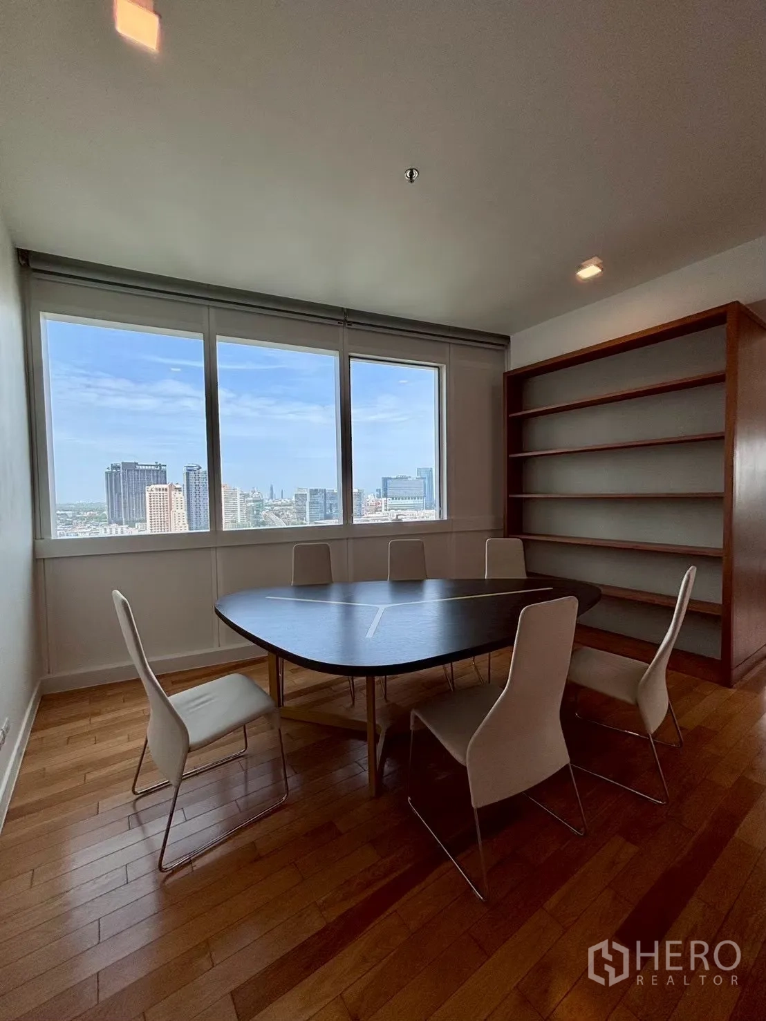 Condominium for rent in Khlong Toei, Bangkok - Dining area with six chairs, large window views of the city, and a tall open bookshelf.