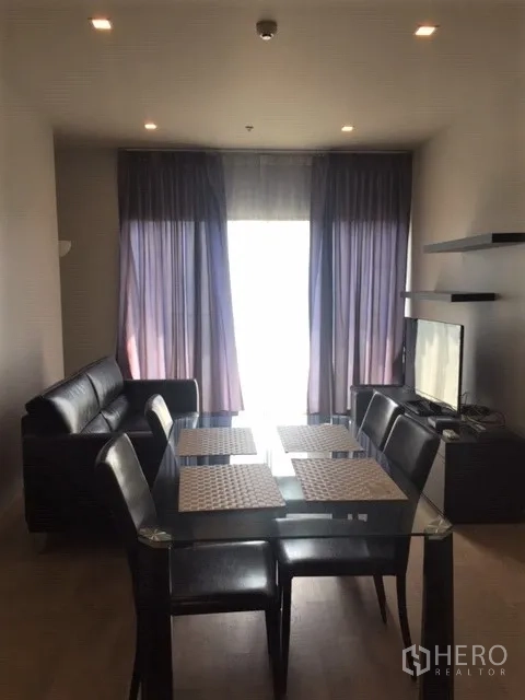 คอนโด ให้เช่าในBangkok - พื้นที่นั่งเล่นและทานอาหารแบบโอเพนแพลน โต๊ะกระจก โซฟาหนัง ทีวี และผ้าม่านสูงจรดเพดาน ในคอนโด 2 ห้องนอนใกล้ BTS พร้อมพงษ์