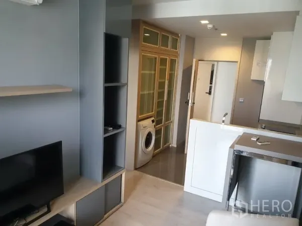 Condominium for rent in Watthana, Bangkok - Modern living room with built-in shelves, TV unit, compact kitchenette, and an in-unit washing machine in the hallway.