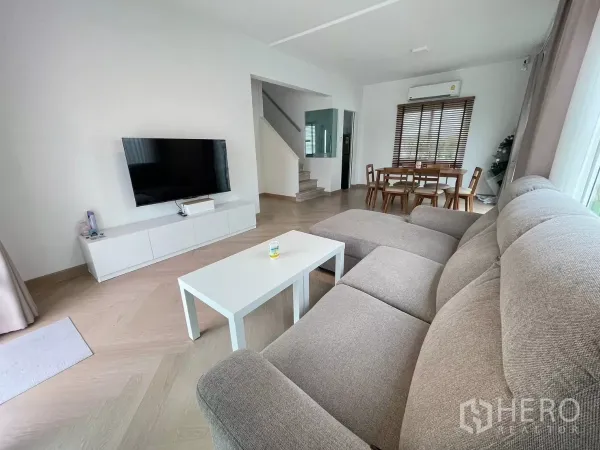 Detached House for rent or sale in Bang Phli, Samut Prakan - Living room with wall-mounted TV, stairs and dining set, showing natural light and chevron flooring.