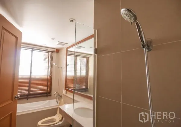 Condominium for rent in Pathum Wan, Bangkok - Bathroom with glass-screened bathtub, handheld shower, vanity and a large window fitted with wooden blinds.