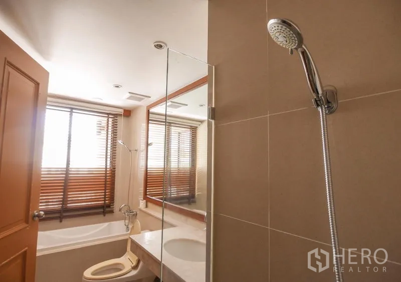 Condominium for rent in Pathum Wan, Bangkok - Bathroom with glass-screened bathtub, handheld shower, vanity and a large window fitted with wooden blinds.