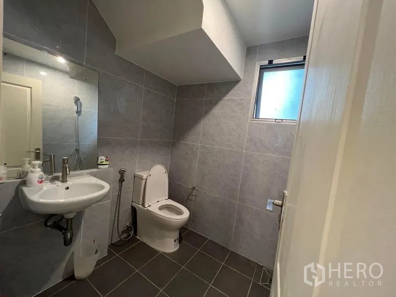 独立式住宅 出售在Bang Kruai, Nonthaburi - 一层卫生间，灰色瓷砖，带窗户、壁挂洗手盆与坐便器。