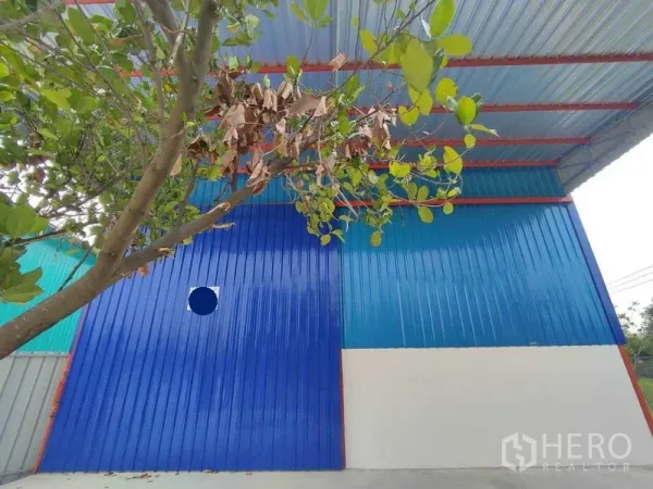 คลังสินค้า ให้เช่าในPak Kret, Nonthaburi - ภาพระยะใกล้ด้านนอก ผนังเมทัลชีทสีน้ำเงินและต้นไม้หน้าตัวอาคาร