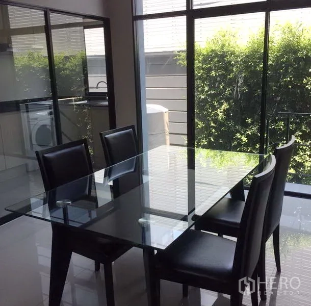 Townhouse for rent in Suan Luang, Bangkok - Bright dining area with a glass-top table and black chairs beside floor‑to‑ceiling windows overlooking greenery.