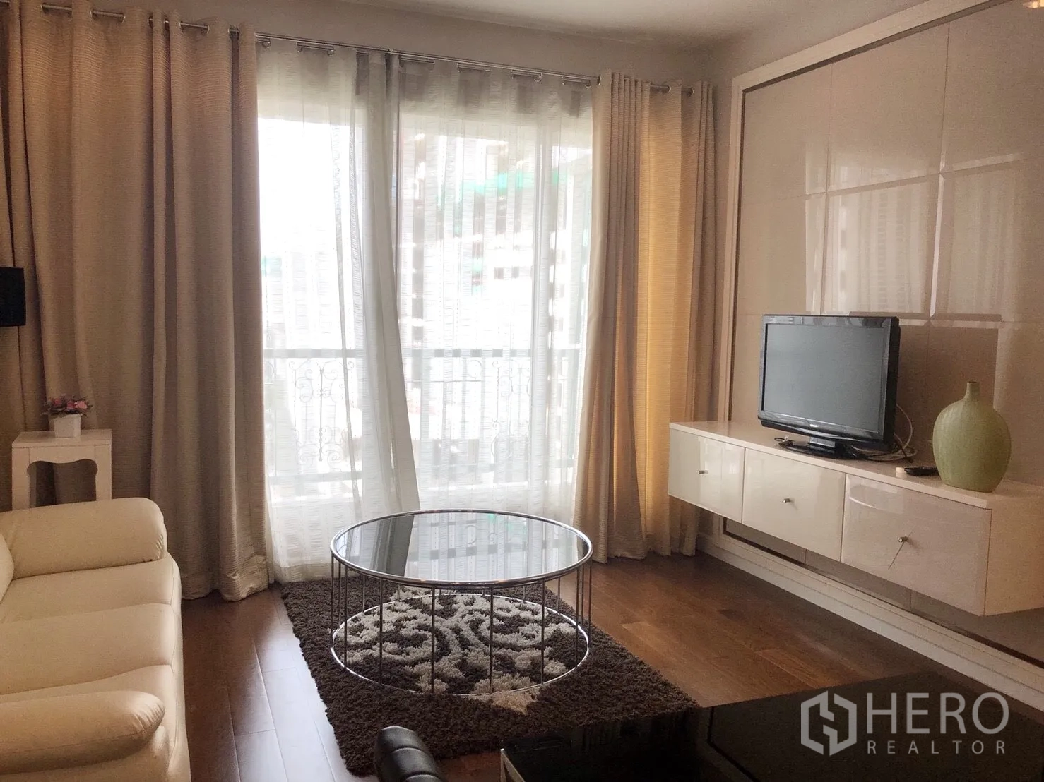 公寓 出租在Pathum Wan, Bangkok - 明亮客厅，落地窗帘与阳台推拉门，白色沙发、圆形玻璃茶几及悬挂式电视柜。