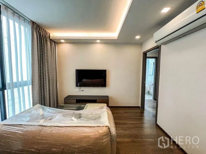 Condominium for sale in Huai Khwang, Bangkok - Bright living room with sofa, wall-mounted TV, air conditioner and doorway to the bedroom.