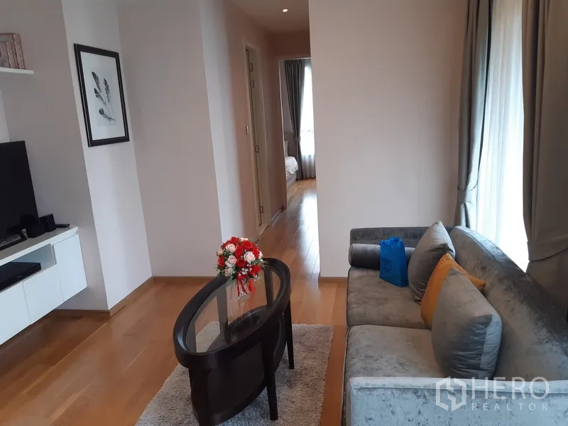 คอนโด ให้เช่าในWatthana, Bangkok - ห้องนั่งเล่นอบอุ่น โซฟาสีเทา ชั้นวางทีวี และทางเดินสู่ห้องนอนบนพื้นไม้