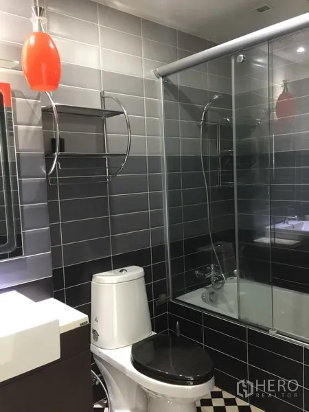 公寓 出租在Watthana, Bangkok - 灰色瓷砖浴室，浴缸带玻璃淋浴隔断与洗手台，橙色吊灯点缀。