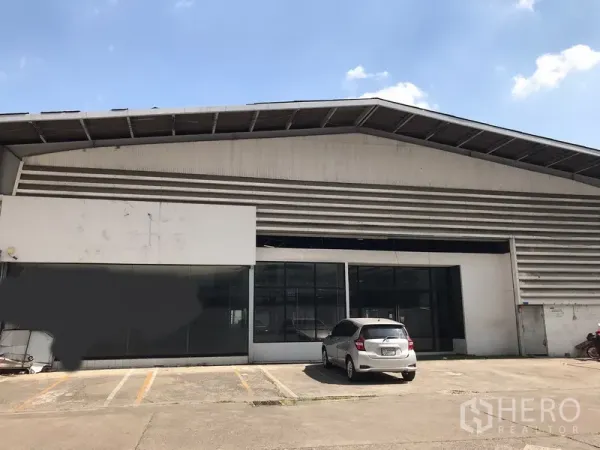Warehouse for rent in Khlong Toei, Bangkok - Front view of a Khlong Toei warehouse with high gable roof, glass-front office and driveway parking under a clear sky.