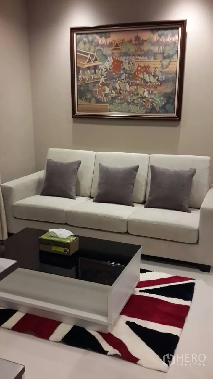 Condominium for rent in Mueang Samut Prakan, Samut Prakan - Bright living room with a cream sofa and gray cushions, glass-top coffee table, and Thai-style wall painting.