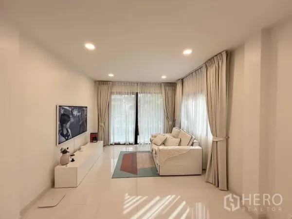 Detached House for sale in Bang Phli, Samut Prakan - Spacious living room with sofa, media wall and floor-to-ceiling curtains opening to the garden.