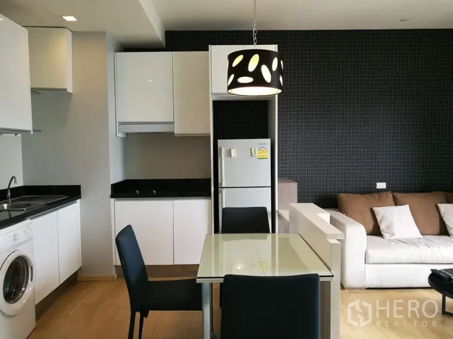 Condominium for rent in Watthana, Bangkok - Dining area with glass table next to a fitted kitchen with washing machine and refrigerator.